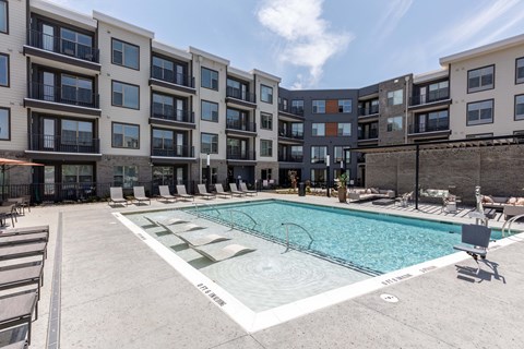 Resort-style pool at Chester Hills with a tanning ledge, in-water loungers, and four-story buildings behind it.