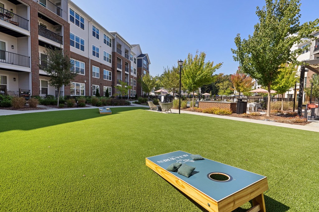 A cornhole board sits on a green lawn in front of apartment buildings.