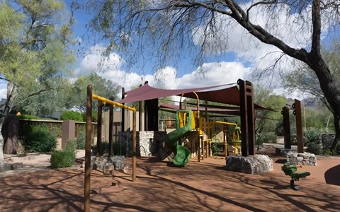 Shaded playground with a green tube slide, yellow climbing bars, swings, and stone walls under mesquite trees.