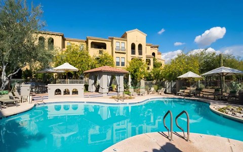 Resort-style pool at Desert Parks Vista with a tile-roofed cabana, shade umbrellas, and stucco buildings behind it.