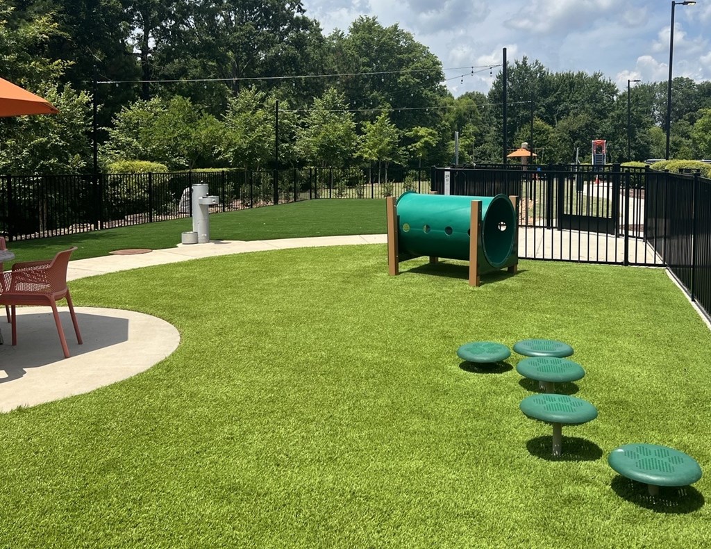 A green cylindrical object is in the middle of a grassy area with a red bench to the left.