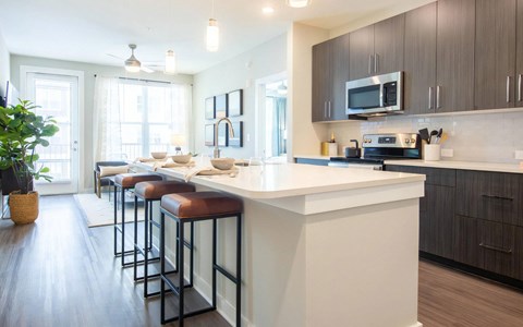 Kitchen island with white quartz, three leather counter stools, dark woodgrain cabinets, and stainless appliances.
