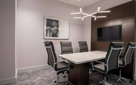 Conference room with a white-topped table, black leather task chairs, a wood panel wall, and a mounted TV screen.