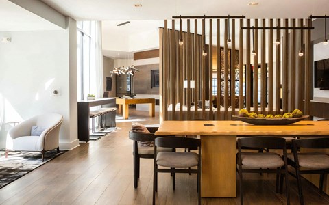 Communal wood table with black chairs under pendant lights, with a bar counter and a pool table in the room beyond.