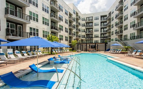 Pool with blue in-water loungers on a shelf, handrails, blue shade umbrellas, and balcony wings behind the deck.