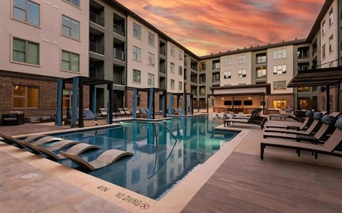 Courtyard swimming pool at sunset with curtained cabanas, rows of chaise loungers, and lit windows in the building.