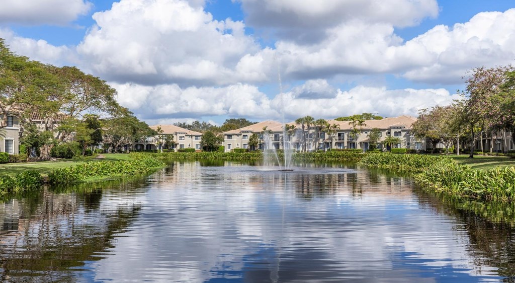 A serene lake with a fountain in the middle surrounded by greenery and houses in the background.