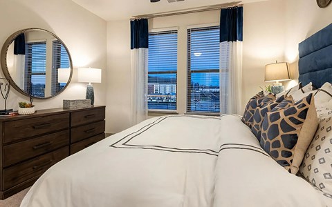 Bedroom with a navy upholstered headboard, two curtained windows, a round mirror, and a wood dresser with a lamp.