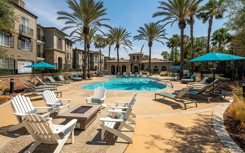 Wide view of the pool deck with a fire pit, Adirondack chairs, chaise lounges, blue umbrellas, and palm trees.