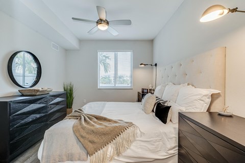 Bedroom with a round black mirror above a textured dresser, a ceiling fan and a shuttered window in the corner.