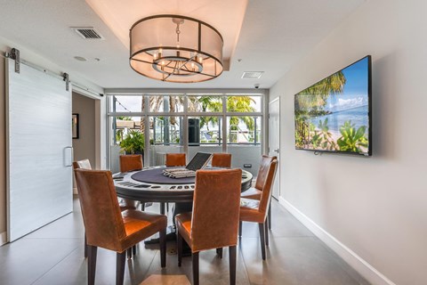 Card room with a round felt game table, eight leather chairs, a drum chandelier and a mounted television on the far wall.