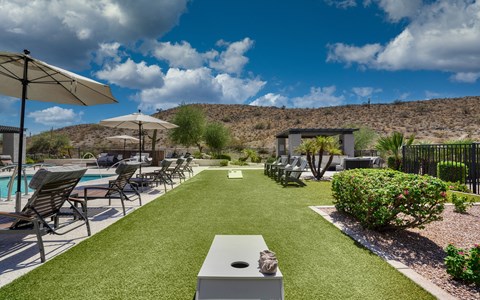 Turf lawn with cornhole boards laid out between the pool deck and a shaded pergola, below the bare desert ridge.