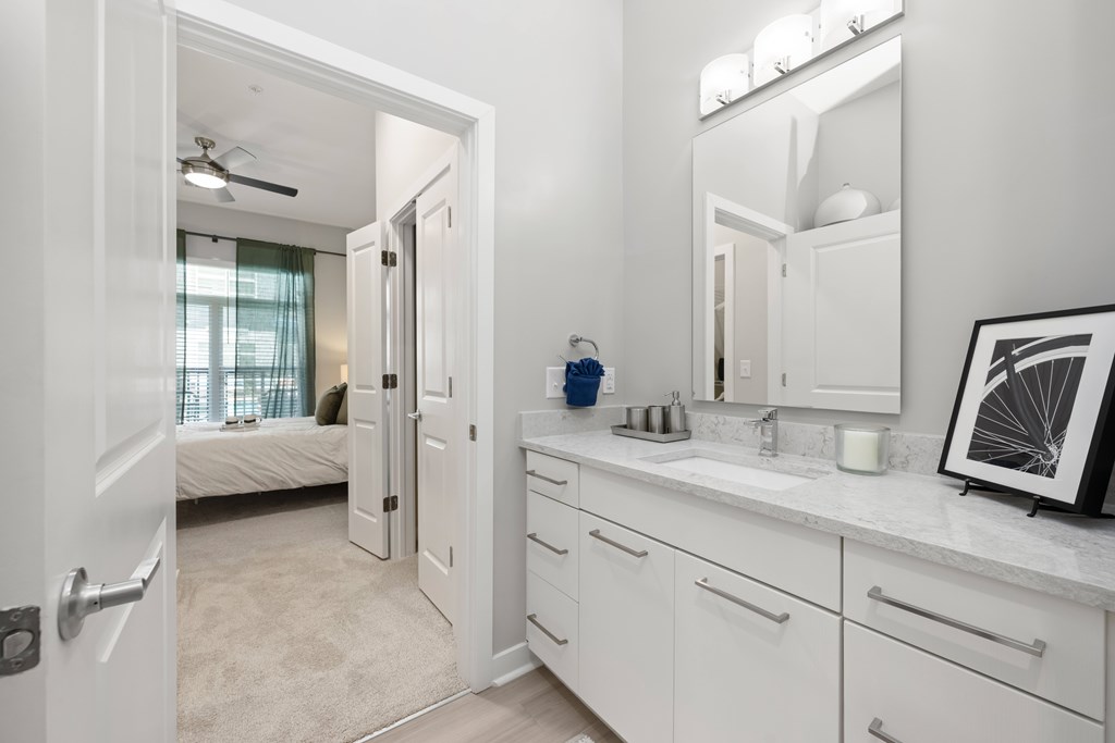 A white bathroom with a mirror, sink, and a bedroom in the background.