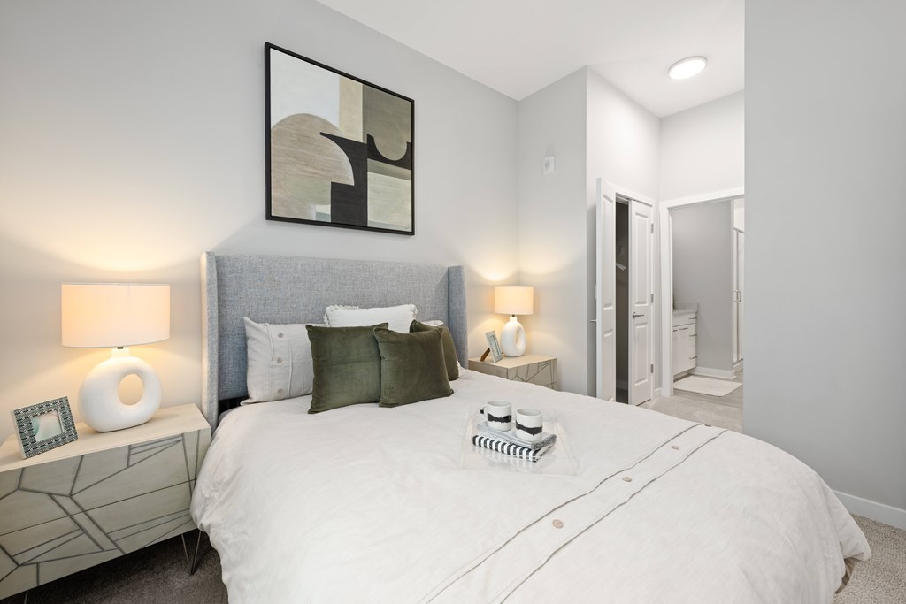 A bedroom with a large bed, a nightstand with a lamp, and a picture frame on the nightstand.