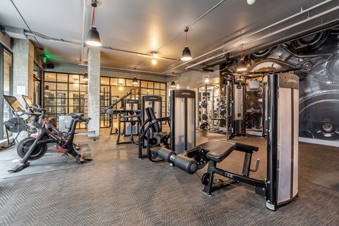 Fitness room with cable machines, benches and cycling bikes, under pendant lights and a large engine wall mural.