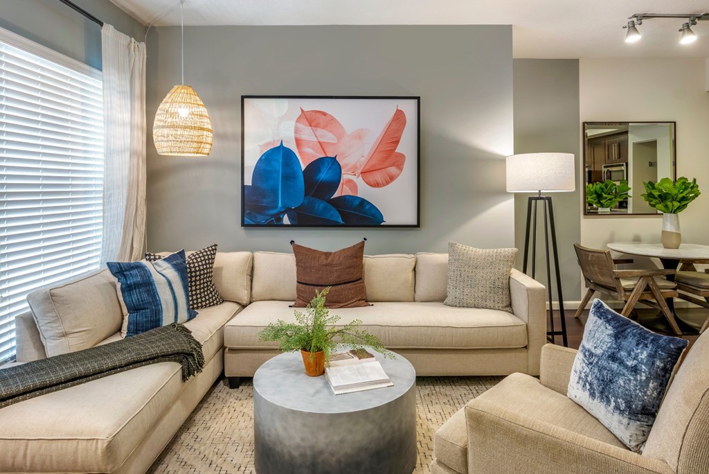 A living room with a large abstract painting on the wall, a beige sofa, a grey coffee table, and a dining table with chairs.
