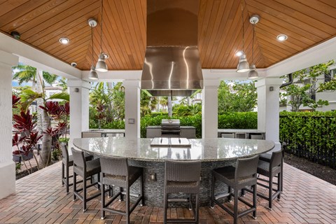Outdoor kitchen under a cedar-ceilinged pavilion, with a curved granite bar, stools, a gas grill and a vent hood.