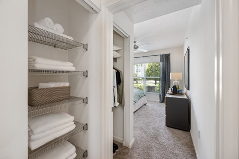 Linen closet with wire shelving stacked with towels, beside an open closet and the carpeted bedroom beyond it.