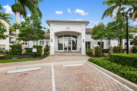 The image shows a white building with a glass entrance and a brick pathway leading to it.