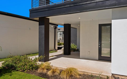 Covered ground-floor patio with a concrete slab, dark columns and planted grasses along the edge of a green lawn.