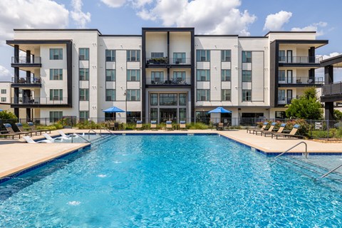 Pool with a wide sundeck of loungers and blue umbrellas, framed by the four-story building on the far side of it.