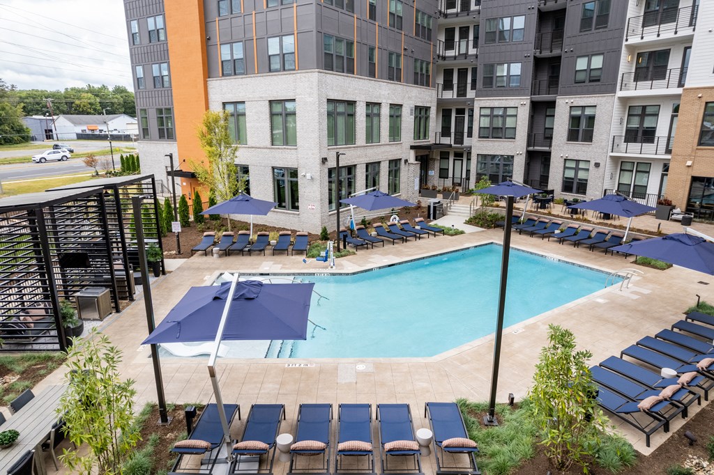 A large swimming pool surrounded by sun loungers and umbrellas in front of apartment buildings.