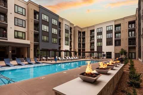 Courtyard pool at sunset with lit fire bowls on a stone ledge, blue chaise lounges, and pergolas along the deck.