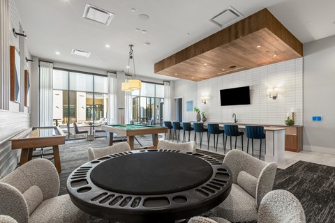 Round poker table with boucle armchairs in the foreground, a shuffleboard, pool table, and bar counter behind.
