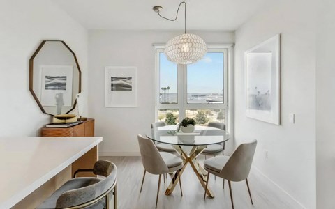 Dining nook with a round glass table on brass legs, upholstered chairs and a beaded pendant over a window onto the water.