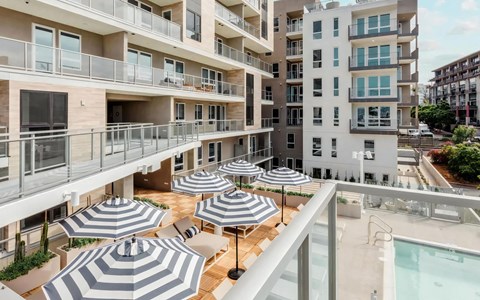 Pool deck seen from a resident balcony, with striped umbrellas, teak decking and the pool along one edge of the frame.
