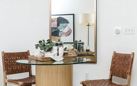 Dining nook with a round glass table on a fluted base, two woven leather chairs and a tall brass mirror on the wall behind.