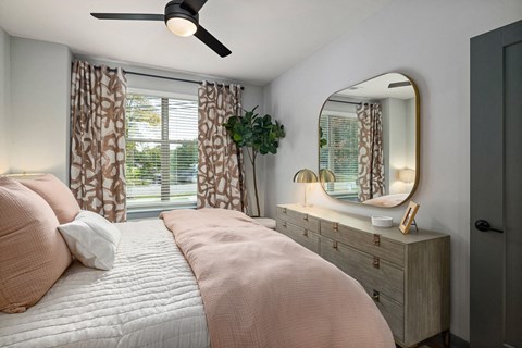 Bedroom with a ceiling fan, patterned curtains, a wood dresser, and a large rounded mirror in a slim gold frame.