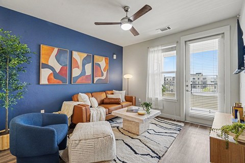 Living room with a blue accent wall, ceiling fan, wood-look floors, and a window beside a glass balcony door with blinds.