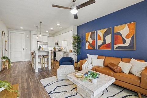 Open living room with a ceiling fan and wood-look floors beside a white kitchen with pendants and an island with stools.