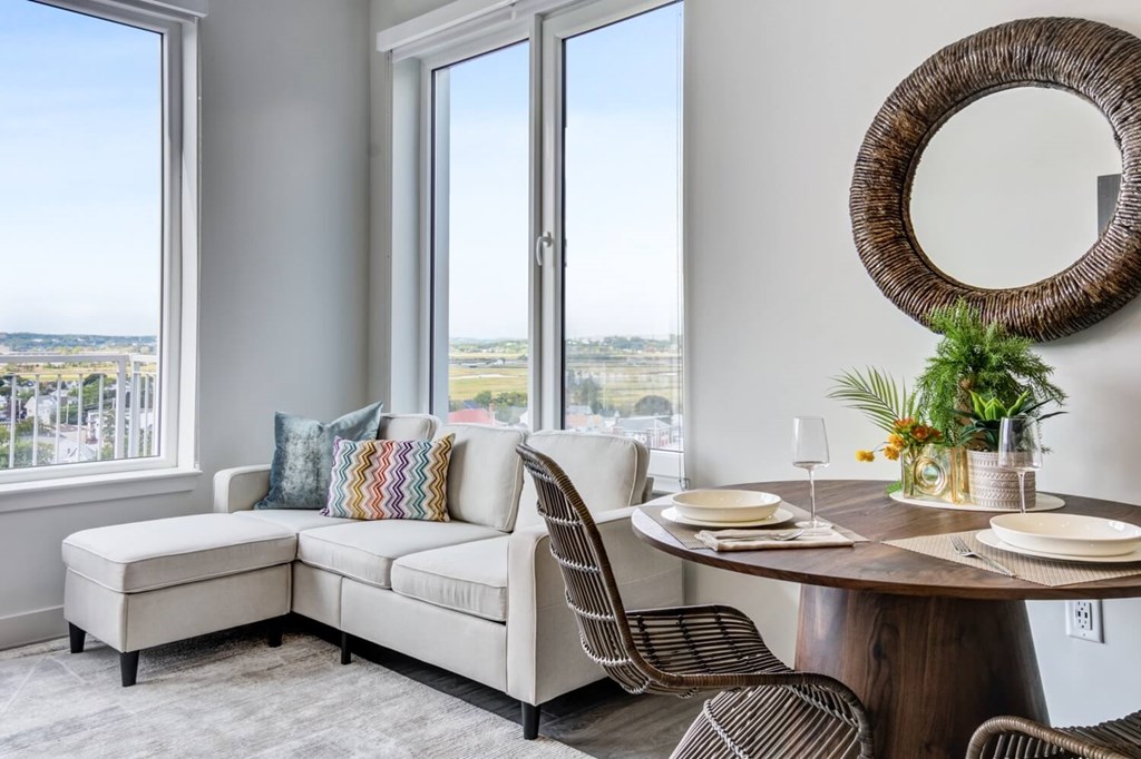 A dining room with a round table and chairs, a bench, and a view of the city through the windows.