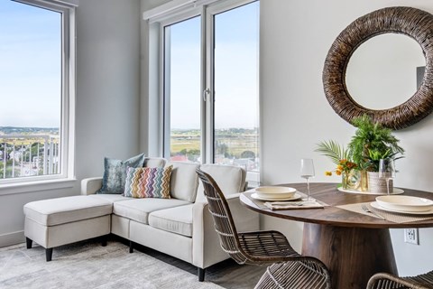 A dining room with a round table and chairs, a bench, and a view of the city through the windows.