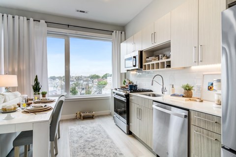 Galley kitchen with pale cabinets, a gas range and quartz counters, beside a dining table set at the wide window.