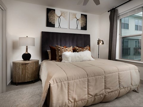 Carpeted bedroom with a tall dark panel headboard, framed botanical art, a round nightstand and a black table lamp.