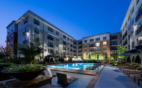 Resort-style pool at Siena Tuscan Village at dusk, with loungers, umbrellas and the lit buildings all around it.