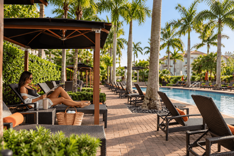 A woman is sitting on a lounge chair by a pool, reading a book.