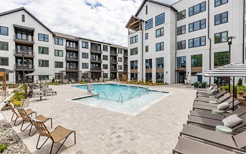 Pool courtyard enclosed by apartment buildings on three sides, with shade umbrellas and long rows of loungers.