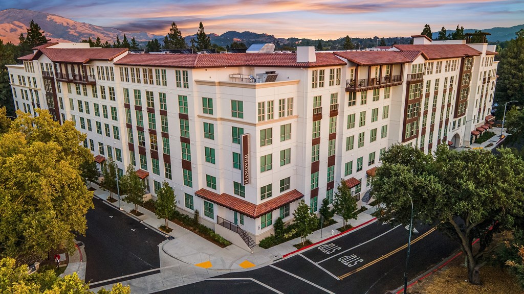 Corner view of a modern apartment exterior with tile rooflines, warm architecture, and tree-lined surroundings