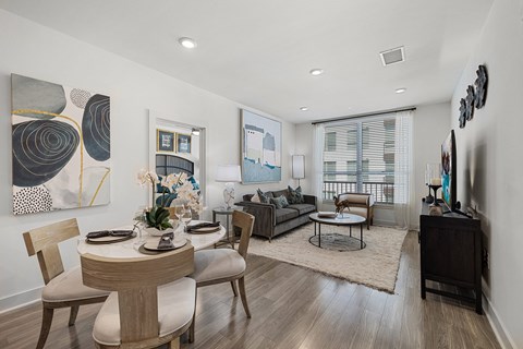 Dining table for four beside a living room with a sofa, a round coffee table and a tall window with roller blinds.