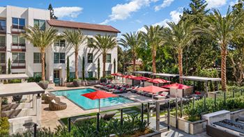 Sunlit pool deck with red umbrellas, cushioned loungers, and shaded cabanas framed by tall palms