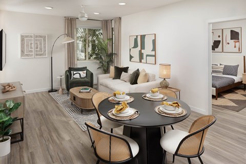 Dining area beside a furnished living room with a bedroom doorway and large windows.