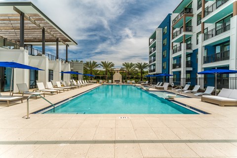 A large swimming pool surrounded by lounge chairs and umbrellas in front of a modern building.