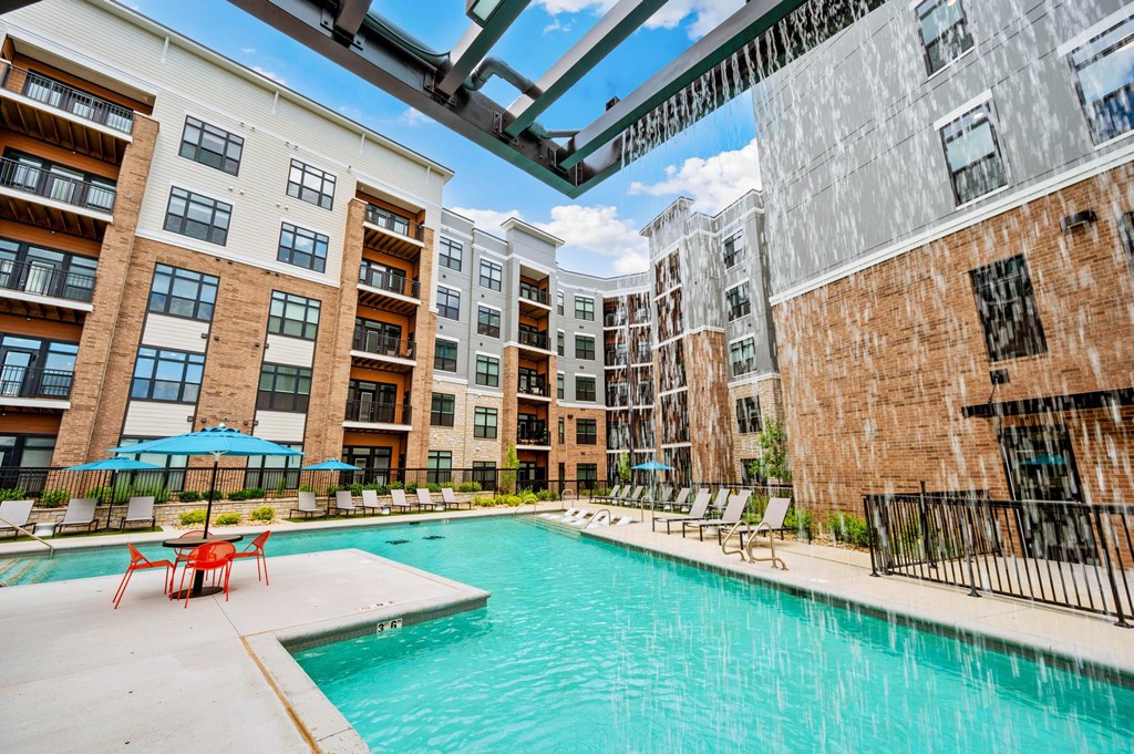 A swimming pool is surrounded by chairs and umbrellas in front of apartment buildings.