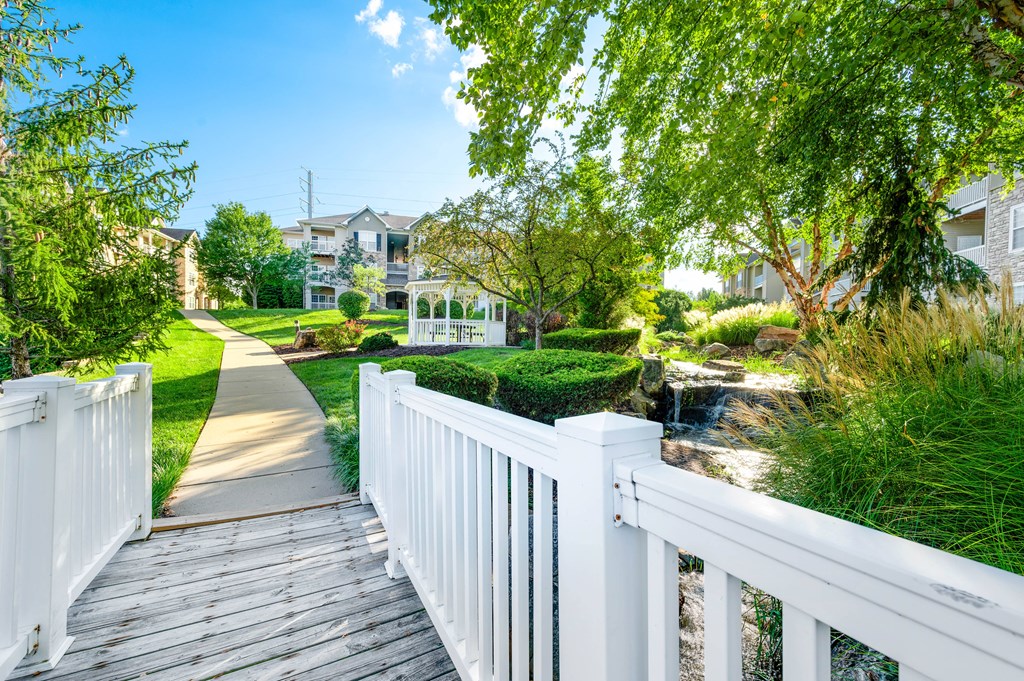 a wooden bridge over a creek with trees on both sides and houses in the background