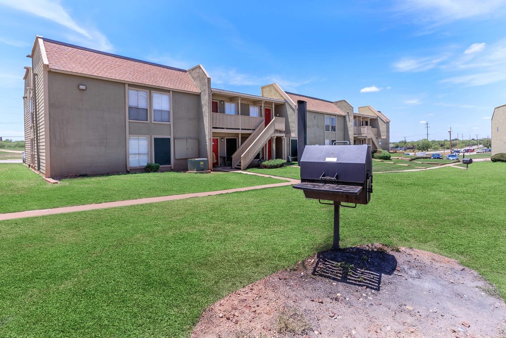 A barbecue grill is in the foreground of a grassy area in front of apartment buildings.