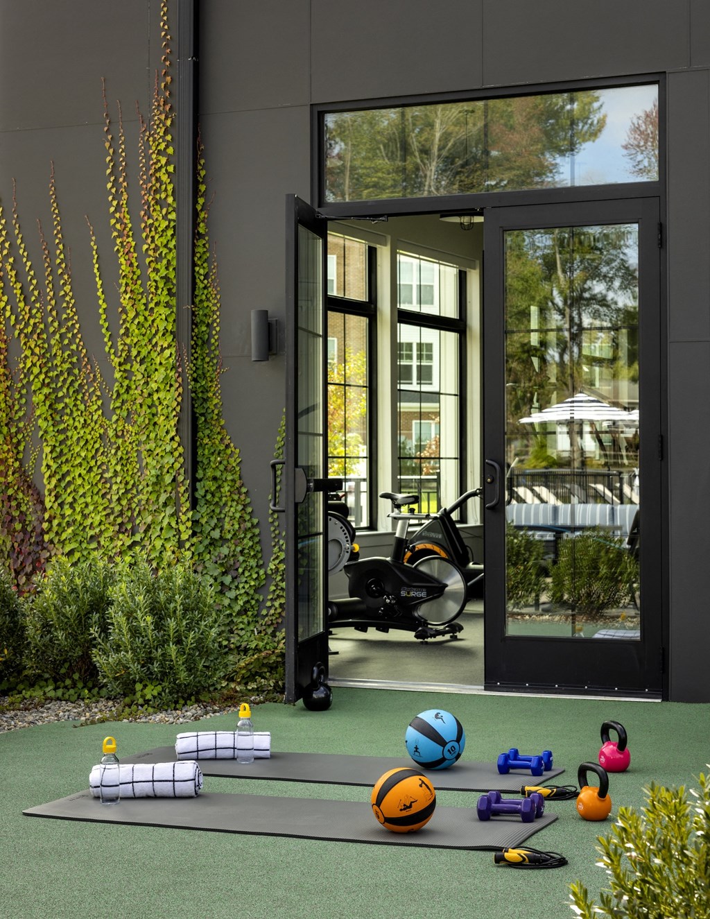 A yoga mat, a foam roller, a stability ball, and a resistance band are all laid out on a green mat outside a modern building.